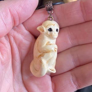May include: Ivory-colored monkey pendant with black eyes and brown accents. The monkey is seated, holding its hands together. The pendant hangs from a black cord with a decorative bronze-colored clasp.