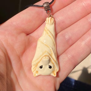 May include: A cream-colored pendant shaped like a bat hanging upside down, featuring a smooth finish with subtle tan shading, black painted eyes, and a detailed snout. The bat is suspended from a silver-toned metal bail on a black cord necklace.