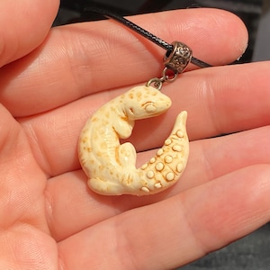 May include: A cream-colored lizard pendant with brown spots and a textured tail, suspended from a black cord by a decorative metal bail. The lizard is curled into a circular shape, showcasing detailed scales and a smooth, carved appearance.