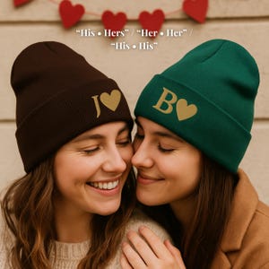 May include: Two beanies, one brown and one green, are shown. The brown beanie has a gold "J" and heart, while the green beanie has a gold "B" and heart. The text "His • Hers" / "Her • Her" / "His • His" is above the beanies.