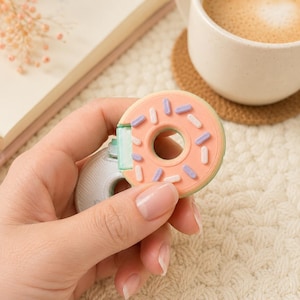 May include: A small, circular, donut-shaped object with a peach-colored top featuring light purple and white sprinkles. The object has a light green hinge mechanism connecting the donut top to a white, textured, semi-circular base component.
