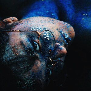May include: A person with blue glitter on their face and body is wearing round glasses. The person's eyes are visible and the glasses are partially covering their face.