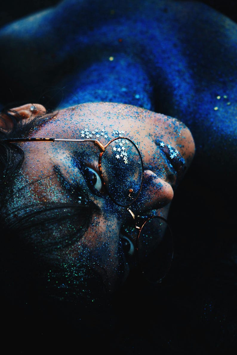 May include: A person with blue glitter on their face and body is wearing round glasses. The person's eyes are visible and the glasses are partially covering their face.