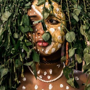 May include: A person with white and gold paint on their face, partially obscured by green foliage. They are wearing a pearl necklace and a black choker.