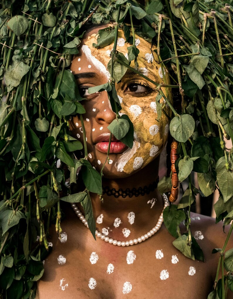 May include: A person with white and gold paint on their face, partially obscured by green foliage. They are wearing a pearl necklace and a black choker.