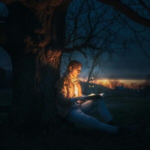 May include: A person sits under a tree at night, reading a book illuminated by a light source. The person is wearing a jacket and jeans. The tree is silhouetted against the night sky.