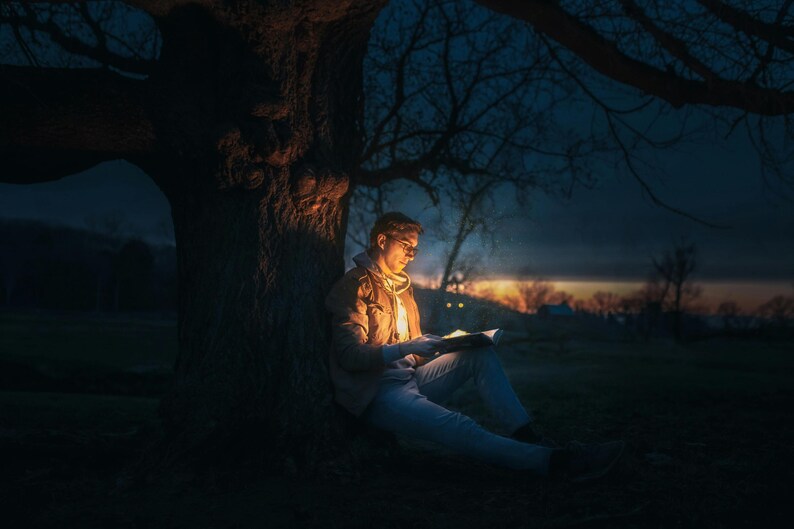 May include: A person sits under a tree at night, reading a book illuminated by a light source. The person is wearing a jacket and jeans. The tree is silhouetted against the night sky.