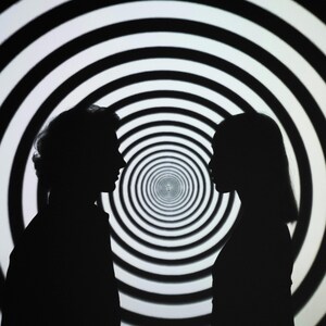 May include: Two silhouettes of people facing each other in front of a black and white swirling pattern.
