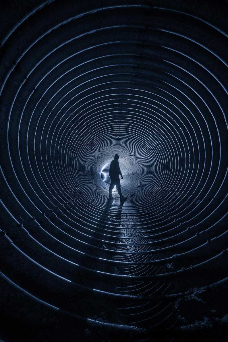 May include: A person stands in the center of a dark, circular tunnel with a bright light at the end. The tunnel is made of metal and has a series of concentric circles.