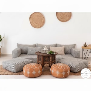 Puede incluir: Un juego de sala de estar con un sofá gris tejido, otomanas cuadradas a juego y dos pufs redondos de cuero. Una pequeña mesa de madera con un juego de té plateado y un plato de verduras se encuentra en el centro. Dos tapices de pared tejidos y una pequeña mesa auxiliar completan la escena.