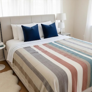 May include: A bed with a beige headboard, white sheets, and two navy blue velvet throw pillows. A striped blanket in shades of beige, gray, red, and blue covers the bed. A small side table with a plant and books is visible.