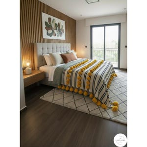 Puede incluir: Interior de dormitorio con una cama con cabecero gris, ropa de cama blanca y cojines decorativos. Una manta a rayas con rayas negras y pompones amarillos está sobre la cama. Una alfombra estampada y paneles de madera completan la decoración.