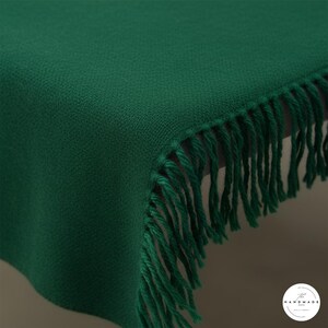 May include: Close-up of a deep emerald green throw blanket with a textured weave. The blanket features a fringe trim along the edge. The handmade blanket is a cozy home decor item.
