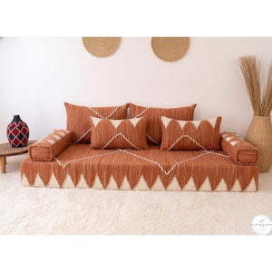 May include: A terracotta-coloured floor cushion set with matching pillows. The set features a geometric design with white accents. Two bolster pillows and four rectangular pillows are included. The set is placed on a white rug.