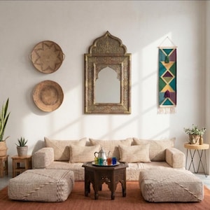 May include: A living room with a beige sofa, two woven ottomans, and a dark wood coffee table. A decorative mirror hangs above the sofa, and woven wall decor is on the wall. A large window lets in natural light, and a red rug covers the floor.