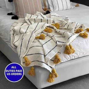 Puede incluir: Una manta blanca con rayas negras y borlas amarillas. La manta está sobre una cama con sábanas y almohadas blancas. El texto "ALL DUTIES PAID US ORDERS" está en un círculo azul.