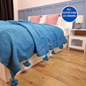 Könnte beinhalten: Eine türkisfarbene Decke mit Quastenbesatz bedeckt ein Bett mit rosa Kissen und einem weißen Kopfteil. Ein weißer Nachttisch mit einer Lampe ist zu sehen. Das Bild enthält einen blauen Kreis mit dem Text "ALL DUTIES PAID US ORDERS".