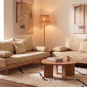 May include: Two beige fabric sofas with wooden frames and matching throw pillows arranged in an L-shape. A round wooden coffee table sits in the center, holding a stack of books and a ceramic mug. A tall floor lamp with a woven shade stands nearby.