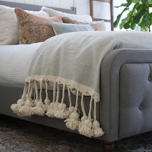Puede incluir: Una cama gris tapizada con una manta gris claro con borlas color crema. En la cama hay cojines decorativos en tonos melocotón, azul y blanco. El marco de la cama está acolchado y tiene una cabecera curva.