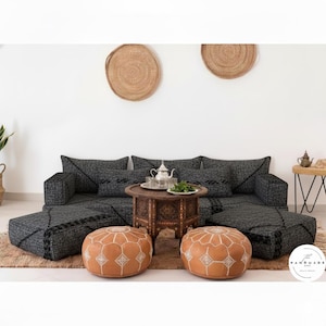 Puede incluir: Un sofá modular gris con ribete negro, combinado con dos otomanas redondas de cuero marrón. Una mesa de centro de madera tallada alberga un juego de té y vegetación. La decoración mural tejida y una pequeña mesa auxiliar completan la escena.