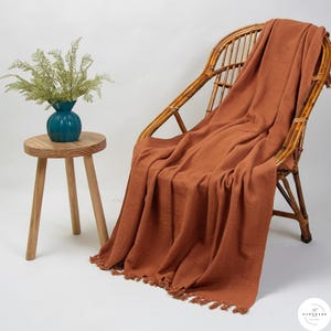 Puede incluir: Una manta de color óxido con flecos está sobre una silla de mimbre. Una pequeña mesa auxiliar de madera sostiene un jarrón turquesa con vegetación. La escena se desarrolla sobre un fondo blanco, creando un ambiente acogedor e invitador.