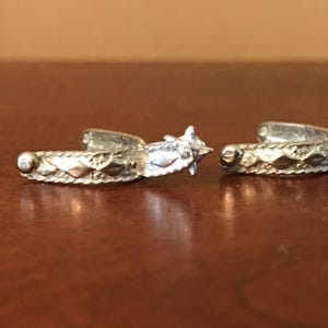 Puede incluir: Un par de gemelos plateados. Cada gemelo presenta un diseño detallado con patrones en forma de diamante y un elemento en forma de estrella. Los gemelos se muestran sobre una superficie marrón.