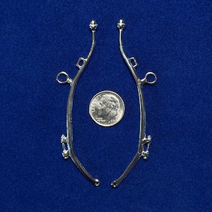 Puede incluir: Dos piezas metálicas plateadas y curvadas con elementos decorativos, posiblemente componentes de joyería, se muestran sobre un fondo azul. Se coloca una moneda de diez centavos estadounidense entre las piezas para comparar el tamaño.