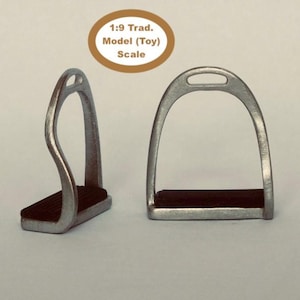 May include: Two silver-colored model stirrups with black footrests. The text on the image reads "1:9 Trad. Model (Toy) Scale". These miniature stirrups are designed for model horses and equestrian displays.