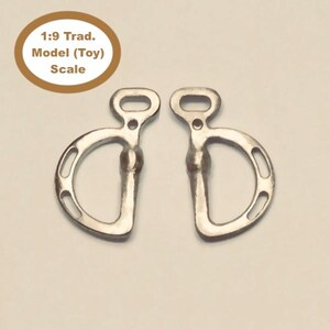 May include: Two silver-colored metal horse bits. Each bit has a D-shaped ring with three holes and an oval-shaped top. The text in the image says "1:9 Trad. Model (Toy) Scale."