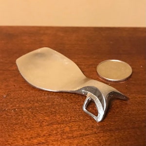 Op de afbeelding: Een zilverkleurig metalen voorwerp met een gebogen, lepelvormige vorm en een kleine, open lus aan het einde. Het voorwerp is ongeveer zo groot als een Nederlandse 20-cent munt.