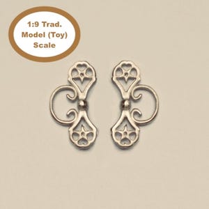 May include: Two silver-toned metal decorative ornaments featuring a symmetrical cutout design with star and heart-shaped motifs, curved side scrolls, and a central sphere. The text 1:9 Trad. Model (Toy) Scale is visible in a brown circular frame.