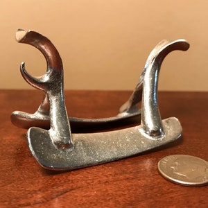 May include: A silver metal stand with two curved arms and a flat base. The stand is about the size of a quarter.