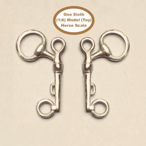 May include: Two silver-colored metal horse bits for a one-sixth scale model toy. Each bit features a large upper ring, a central shank, and a smaller lower ring. The text "One Sixth (1:6) Model (Toy) Horse Scale" is printed in the center above the bits.