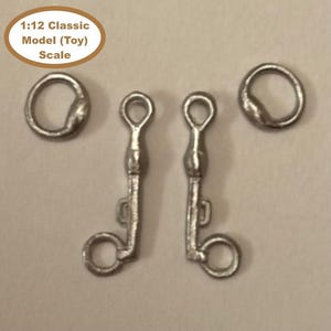 May include: Miniature silver-coloured metal horse tack set, including two bit rings and two bit shanks. The text "1:12 Classic Model (Toy) Scale" is in a brown oval in the upper left corner. The set is designed for a toy horse.