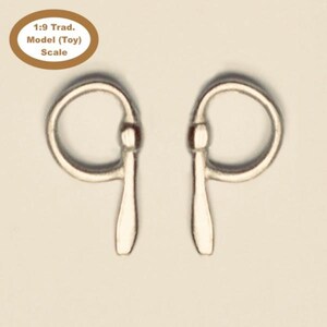 May include: Two silver-coloured metal horse bits. Each bit has a circular ring on top, connected to a vertical bar with a teardrop-shaped end. The text "1:9 Trad. Model (Toy) Scale" is in a brown oval in the upper left corner.