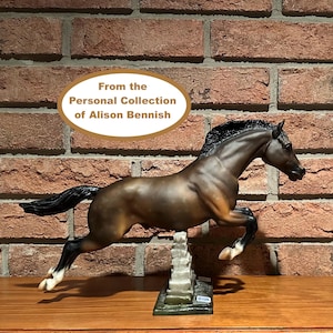 Puede incluir: Figura de caballo marrón y negro en pleno salto, montada sobre una base decorativa. La figura tiene un acabado brillante y detalles precisos. El texto "From the Personal Collection of Alison Bennish" se muestra encima del caballo.
