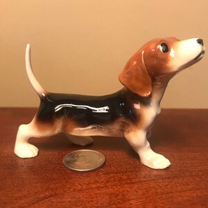 May include: A ceramic figurine of a beagle dog with brown, black, and white markings. The dog is standing on all four legs with its head tilted up and its tail curled.