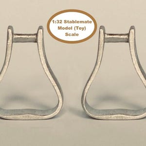 Puede incluir: Dos estribos plateados, equipo ecuestre, se muestran sobre un fondo neutro. El texto "1:32 Stablemate Model (Toy) Scale" está en un óvalo encima de los estribos. Los estribos son de metal.
