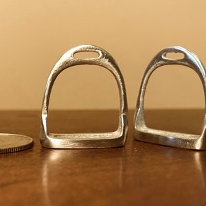 Puede incluir: Dos llaveros de metal plateado en forma de estribo. Los llaveros son aproximadamente del tamaño de una moneda de 1 euro.