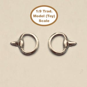 Puede incluir: Dos anillos de metal plateado, cada uno con una pequeña protuberancia puntiaguda en el borde exterior. Sobre los anillos, un texto en un óvalo marrón dice 1:9 Trad. Model (Toy) Scale. Estos artículos son para figuras a escala 1:9.