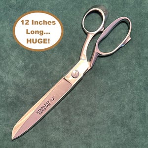 May include: Large stainless steel scissors measuring 12 inches in length. The blades are engraved with the text 'STAINLESS PAKISTAN 12"'. A circular graphic in the upper left corner contains the text '12 Inches Long... HUGE!'.