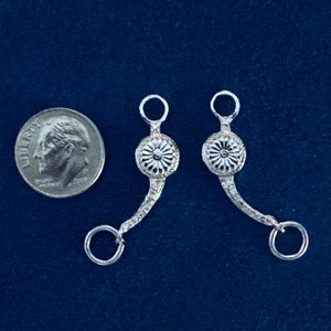 May include: Two silver-toned metal horse bit pieces with a decorative floral design. The bit pieces are about the size of a UK ten pence coin.
