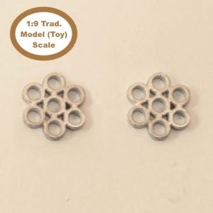 May include: Two silver-colored, flower-shaped toy model parts. Each part has a central design of interlocking circles surrounded by a ring of circular holes. The text "1:9 Trad. Model (Toy) Scale" is in a circular label.