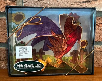 Könnte beinhalten: Ein goldener Hähnchen-Ausstecher in einer durchsichtigen Plastikbox mit blauem und grünem Hintergrund. Die Box ist mit einem goldenen Band verziert. Der Text auf der Box lautet "BOOTH #103 Item #02-005 Rooster Copper Cookie Cutter 9,99 $ Ann Clark Ltd. The Cookie Cutter People From Vermont."