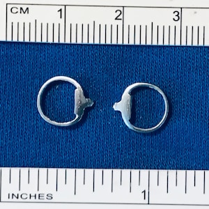 May include: Two small, silver-colored metal components, each with a circular ring and a rectangular tab with a small protrusion. The components are on a blue fabric surface, with a ruler showing inches and centimeters.