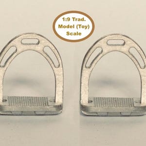 Puede incluir: Dos estribos de juguete plateados para caballos. Cada estribo tiene una parte superior arqueada con tres recortes ovalados y un reposapiés rectangular con ranuras horizontales. El texto "1:9 Trad. Model (Toy) Scale" está en un círculo marrón.