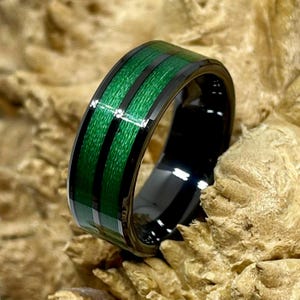 May include: A black ring with a green and black striped design. The ring has a glossy finish and is set against a textured, light brown background. The ring's band features a series of vertical green stripes separated by black lines.