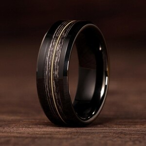 May include: A black tungsten carbide ring with a wood inlay and a thin gold band. The ring has a beveled edge and a polished finish.