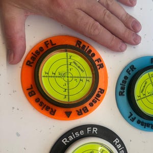 Puede incluir: Tres niveles circulares en naranja, azul y negro. Cada nivel tiene un centro amarillo brillante con un indicador de burbuja y el texto "Raise" y "FL", "FR", "BR", y "BL". Estos niveles están diseñados para tareas de nivelación y alineación.