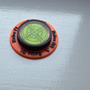 May include: A circular orange 3D-printed base with a black rim and a central green bullseye spirit level. The orange base features black embossed text reading Raise FL, Raise BR, and Raise BL with directional arrows for levelling adjustments.